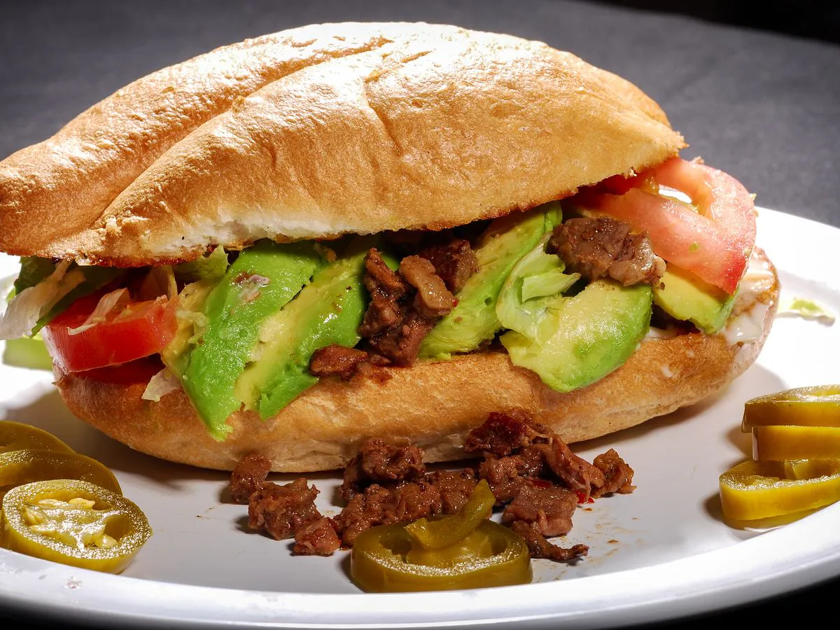 Crusty sandwich with avocado, tomato, and meat, served with pickled jalapeños on a plate